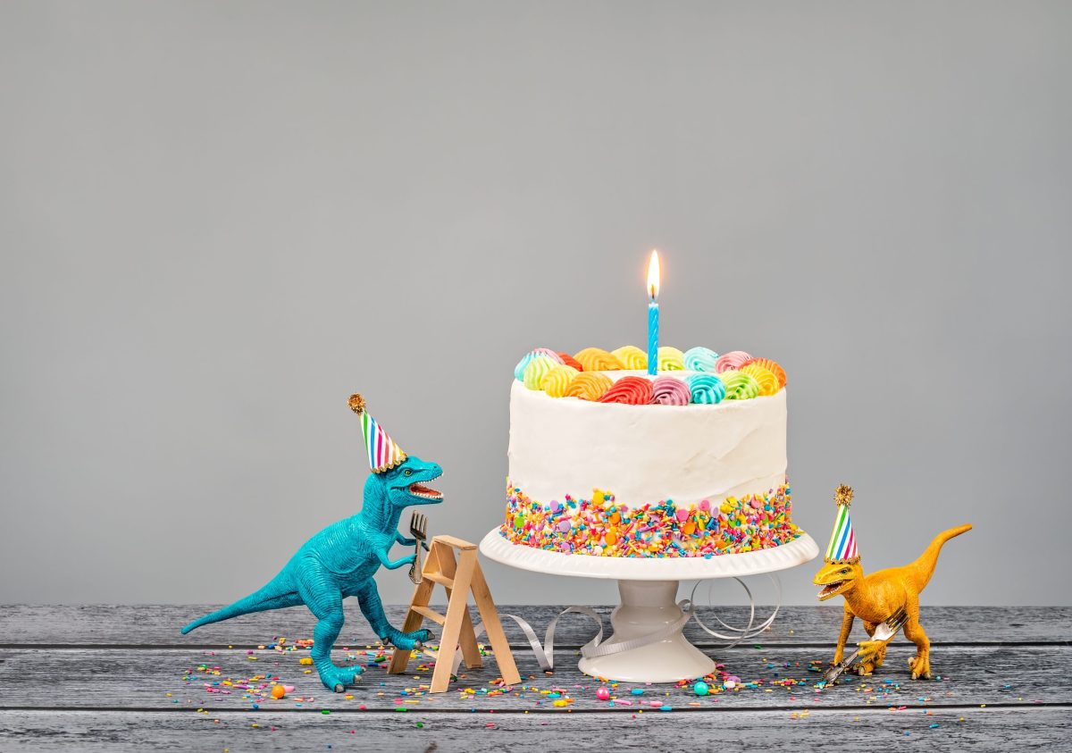 Hungry toy dinosaurs wearing hats and holding forks next to a birthday Cake on a gray background